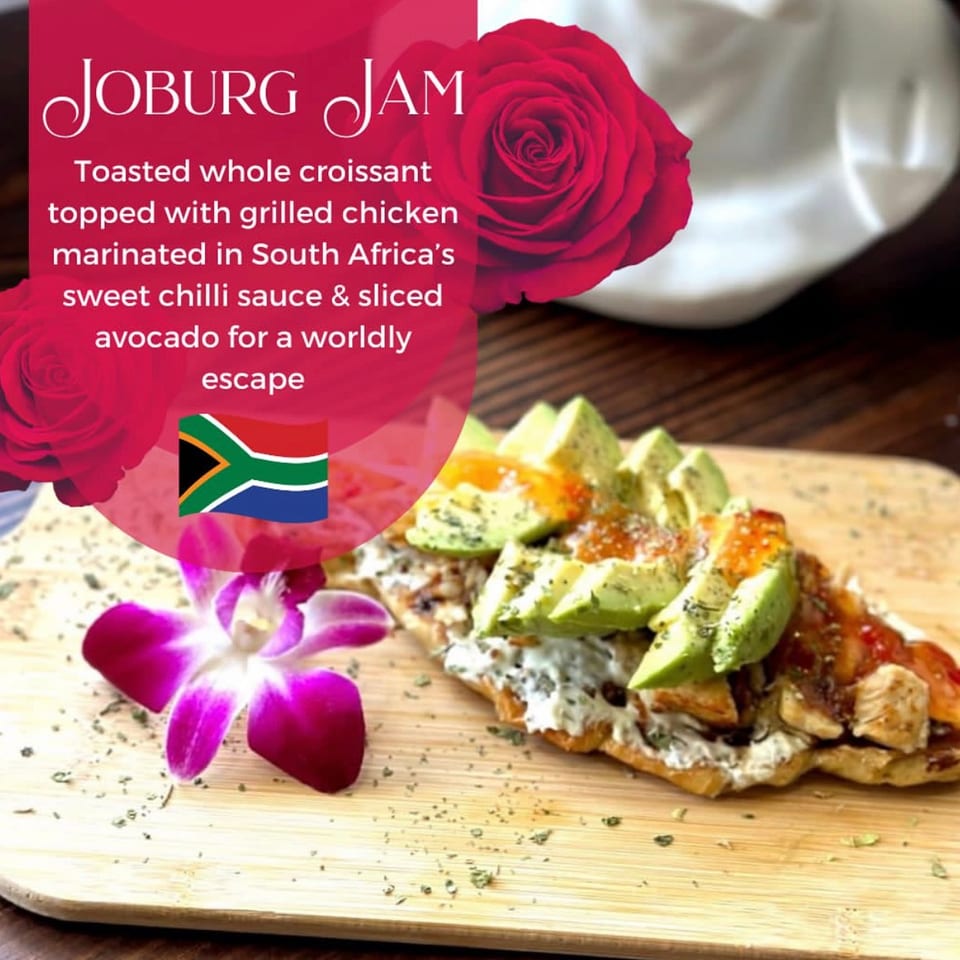 Toasted croissant topped with grilled sweet-chili chicken, sliced avocado, creamy sauce, and edible flowers on a wooden board.