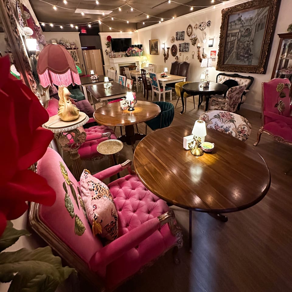 Eclectic restaurant dining room with pink velvet seating, wooden tables, vintage artwork, floral decor, and warm string lights.