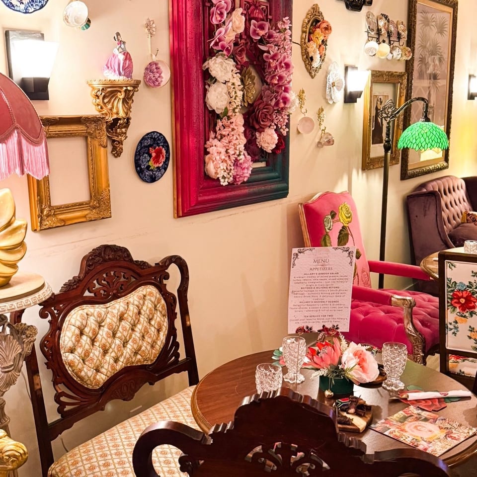 Eclectic restaurant dining room with floral artwork, ornate vintage chairs, a set table, pink seating, and a Tiffany-style lamp.