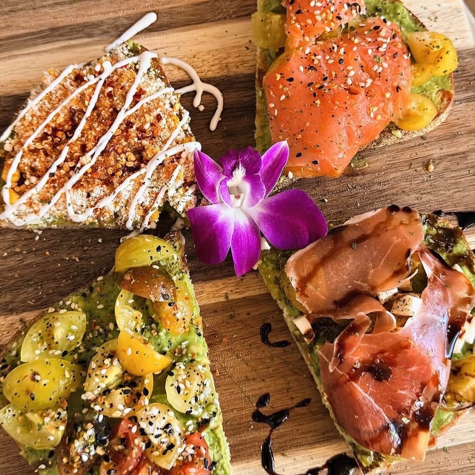 Wooden platter of avocado toast topped with salmon, prosciutto, tomatoes, seeds, sauces, and a purple orchid garnish.