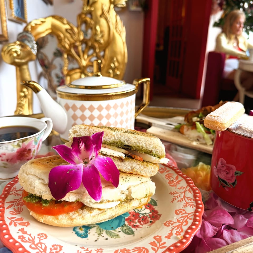 Afternoon tea spread with floral sandwiches, coffee, red drink, and ornate gold decor in an elegant dining room.