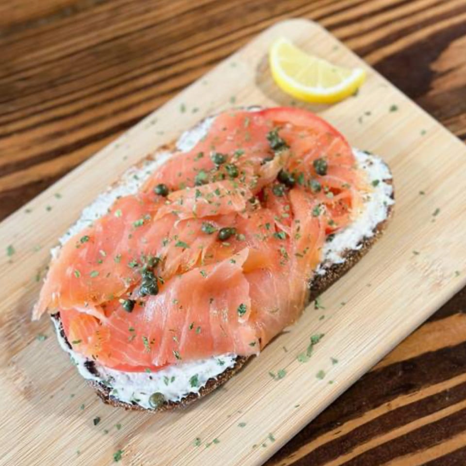 Open-faced smoked salmon toast with cream cheese, capers, herbs, and lemon wedge on a wooden board.